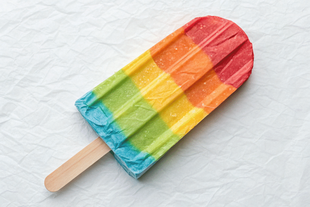 A close-up photograph of a vibrant, multi-colored paper squishy ice pop with a variety of colors layered together, creating a visually striking appearance. The colors include shades of red, orange, yellow, and green. The ice pop is positioned against a neutral, light-colored background.