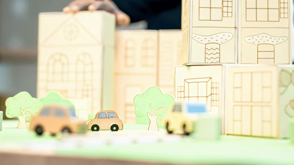 A photo of a creative and playful scene featuring a table with a miniature toy cityscape and toy cars The scene is composed of several cardboard blocks with various structures and illustrations including buildings with windows green foliage and a simplistic cartoonish design