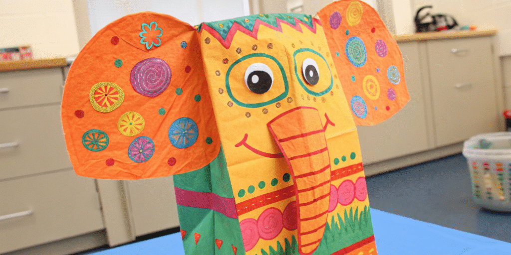 Orange elephant paper bag puppet simply paper crafts The image shows a colorful elephant paper bag puppet decorated with various vibrant colors and patterns created by children using crayons and stickers The puppet features a long accordion folded trunk and large floppy ears showcasing the kids' creativity and fine motor skills in a fun craft project.