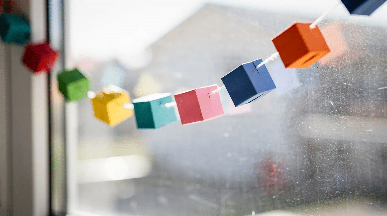 A colorful garland made of origami paper cubes hangs gracefully against a bright window showcasing a vibrant display of various shades Each finished cube crafted from square sheets of paper adds a playful touch to the room making it a delightful decoration for school projects or DIY crafts