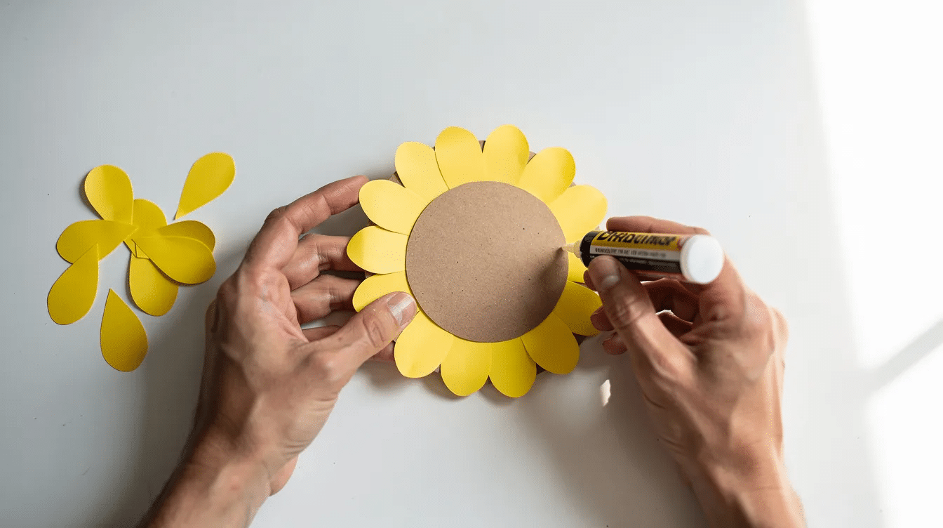 66458e84 f3c6 4f43 a975 cc943d2abf16 A pair of hands is gluing bright yellow paper petals around a central brown circle creating a vibrant paper sunflower craft This step in the paper flower tutorial showcases the fun of making colorful crafts that can brighten up any space