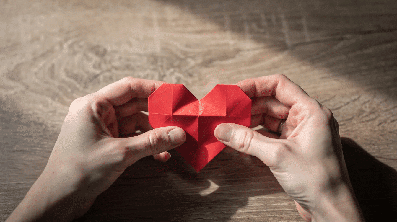 F643947f fa33 4428 b403 de3dccd8cd68 Two hands are carefully folding a small red origami heart from a square piece of paper on a wooden table showcasing the intricate process of creating a traditional origami heart The left hand holds the bottom edge while the right hand aligns the top points demonstrating the step by step instructions to make a paper heart